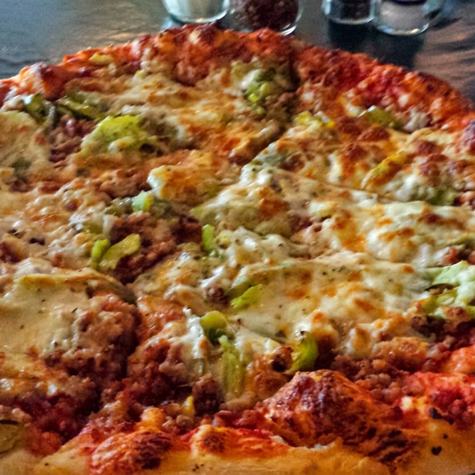 Close-up of a loaded pizza topped with melted cheese, green peppers, and crumbled meat on a dark restaurant table.